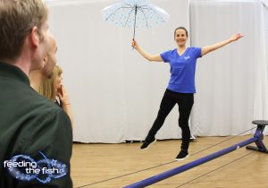 A woman walks a tightrope and holds an umbrella as three people watch