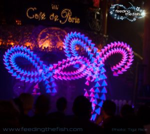 Two performers on stage at Café de Paris spin LED light wands. They display graphics of ribbons tied in a bow, in blue and pink.