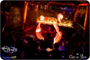 Two performers on stage at Café de Paris spin LED light wands. They display orange graphics in the air.