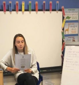 A female teacher reads from a piece of paper in a school classroom