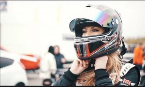 A female stunt driver unfastens her helmet as she looks outwards proudly.