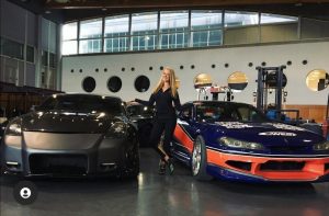 A blonde woman stands and smiles between two race cars.