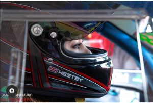 A woman wearing a driving helmet sits in a sports car, she looks out of the visor and the photo is taken from a profile angle.