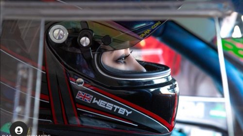 A woman wearing a driving helmet sits in a sports car, she looks out of the visor and the photo is taken from a profile angle.